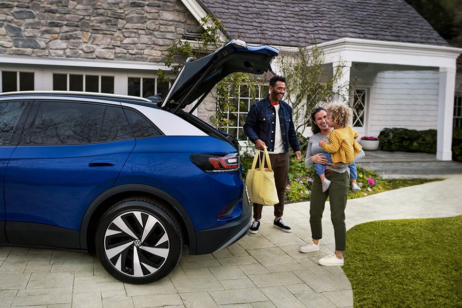 A young family loads a bag into the cargo area of an ID.4, shown in Dusk Blue Metallic parked in a residential home’s driveway.